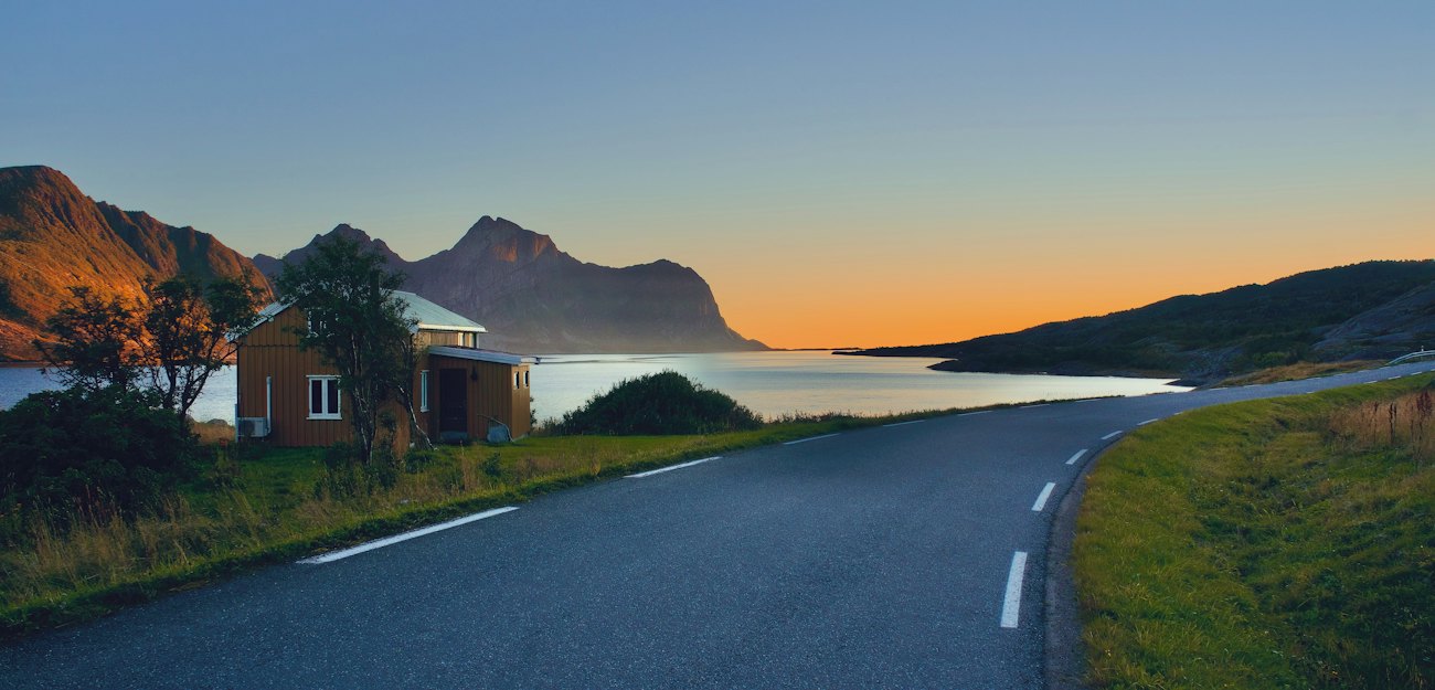 An empty coastal road curving past a cabin toward mountains under an orange midnight-sun sky