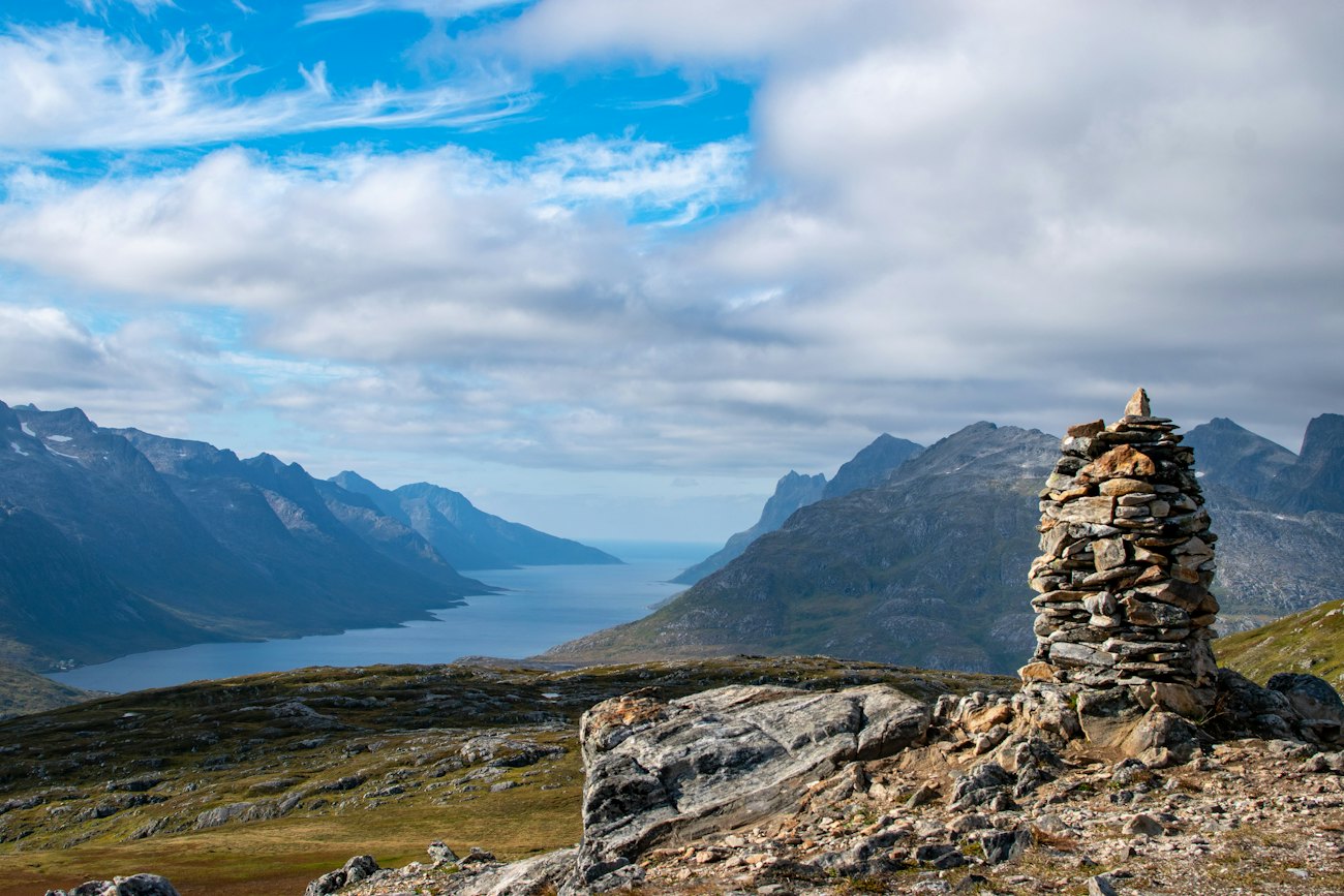 A stone cairn on a mountain top overlooking a fjord and distant peaks