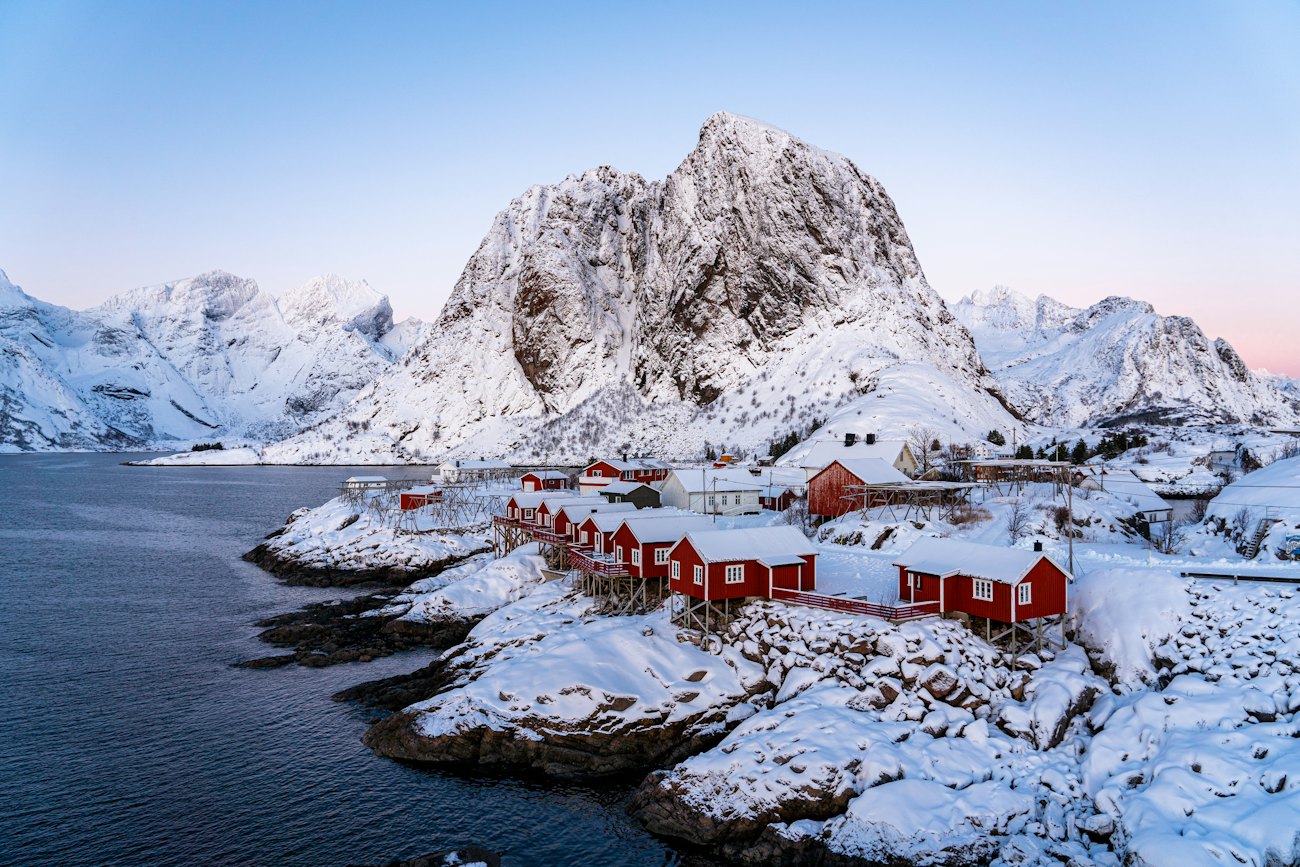 Red fishing cabins on snow-covered rocks beneath a sharp white peak at Hamnøy in Lofoten
