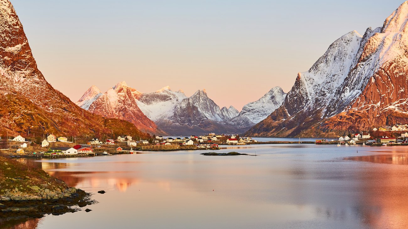 Pink morning light on snowy peaks above the still water at Reine in Lofoten