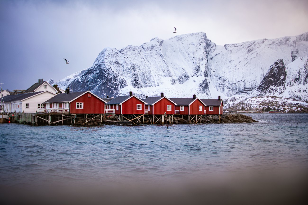 A row of red rorbu cabins on stilts over the water with a snowy mountain behind