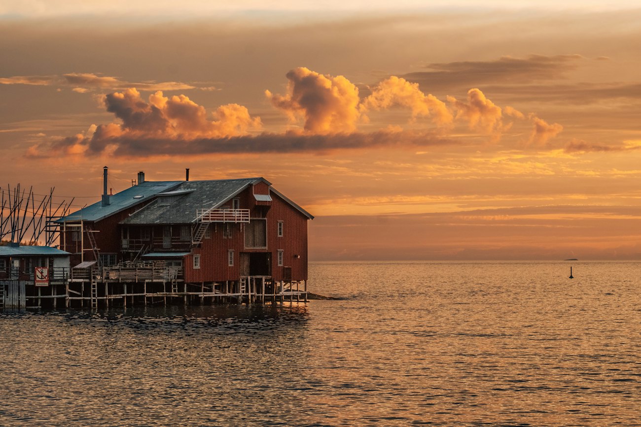 A red wooden cabin on stilts over calm water under an orange sky