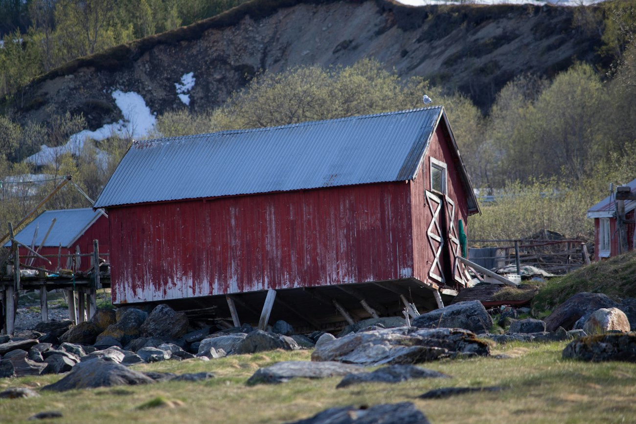 A weathered red boathouse on a rocky shore beneath a mountain