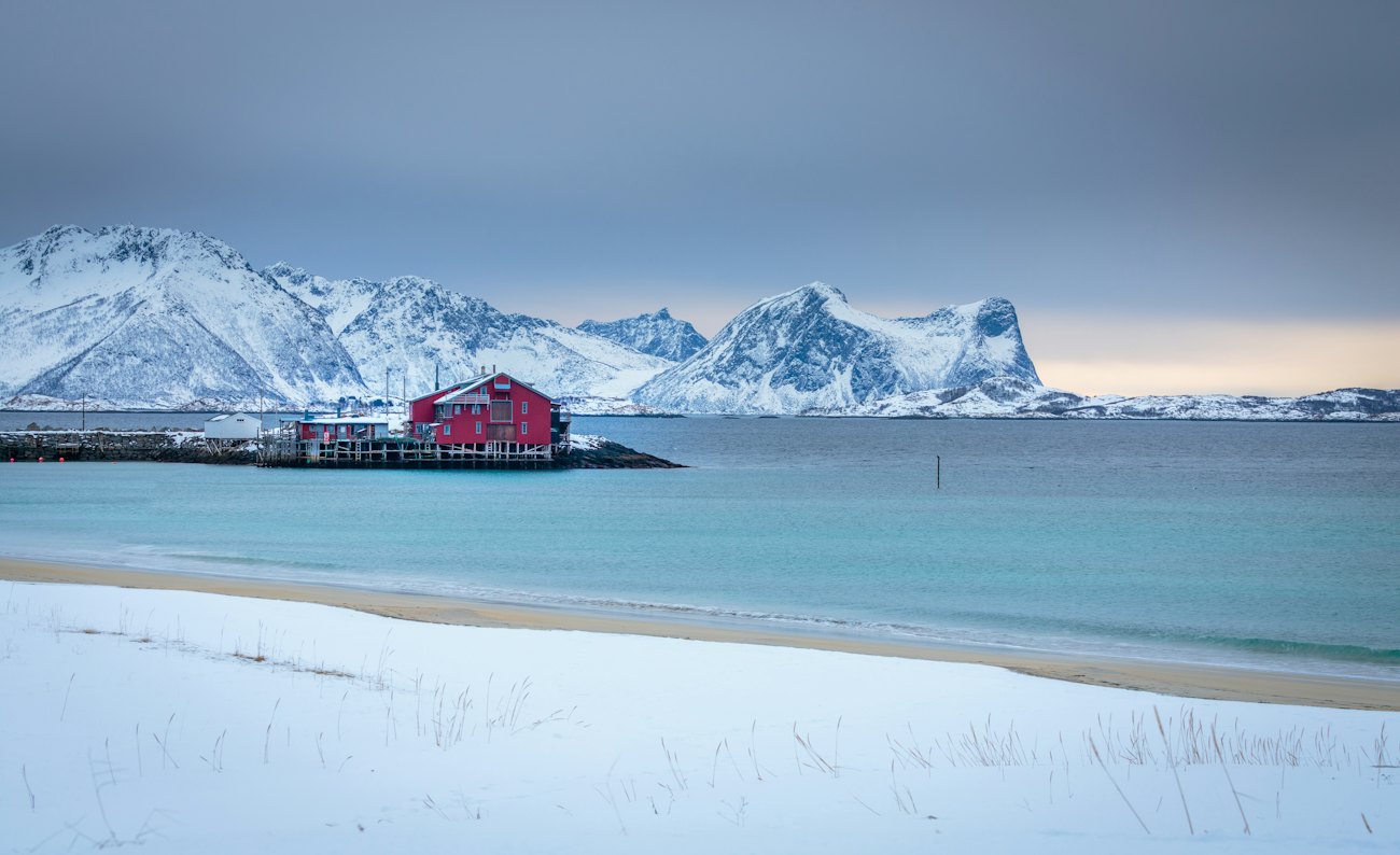 A red fishing cabin on a pier beside a snowy beach and steep white mountains